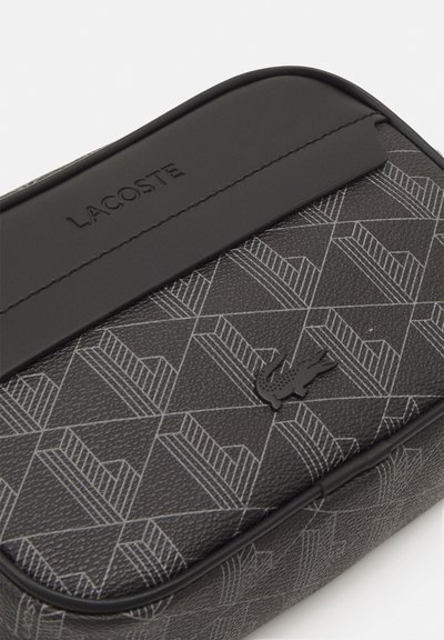 Pochette noire Lacoste avec un motif géométrique, extérieur texturé, logo en relief et panneau supérieur noir lisse, avec des coutures contrastées et un emblème.