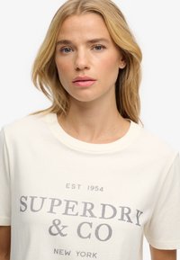 Cream-colored t-shirt featuring a crew neck, short sleeves, and "SUPERDRY & CO" printed in dark gray text. Soft cotton material.