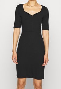 Robe noire ajustée avec un col festonné, des manches courtes et une texture lisse, se terminant juste au-dessus du genou. Design minimaliste sans motifs.
