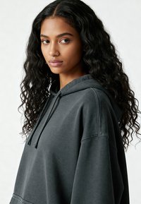 Giovane donna con lunghi capelli neri ricci, che indossa un ampio hoodie grigio scuro, che guarda leggermente di lato su uno sfondo chiaro.