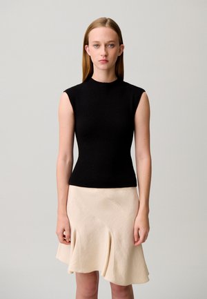 Giovane donna con capelli lisci castano chiaro, che indossa un top nero senza maniche e una gonna svasata color crema, in piedi contro uno sfondo semplice.