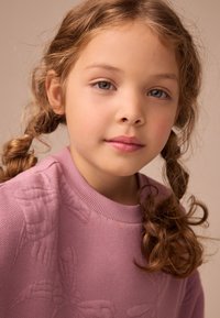 Lyserød strikket sweater med teksturerede blomstermønstre, med rund halsudskæring og ribstrikkede ærmegab. Blødt, bølget hår stylet i to fletninger.