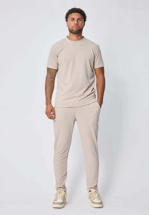 Homme tatoué debout, portant une chemise beige à manches courtes, un pantalon assorti et des baskets beiges et blanches sur un fond clair uni.