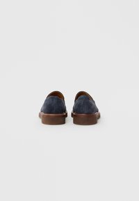 Egy pár sötétkék velúr slip-on cipő barna bőrsarokkal, hátulról egy fehér háttér előtt.