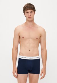 Boxers en coton bleu marine avec une ceinture élastique blanche arborant le logo "LACOSTE". Texture lisse, design ajusté, longueur jusqu'au mi-cuisse.