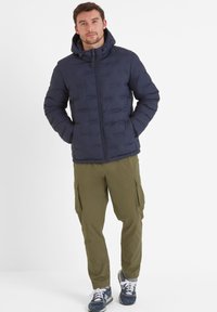Navy padded jacket with a hood, featuring a front zipper and side pockets, paired with olive green cargo pants and blue athletic shoes.