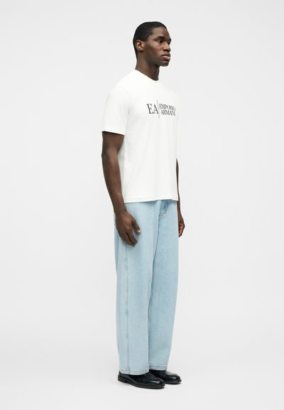 Άνδρας που φοράει λευκό T-shirt Emporio Armani, ανοιχτό μπλε τζιν παντελόνι φαρδύ, και μαύρα παπούτσια, στέκεται πλαγίως πάνω σε απλό λευκό φόντο.