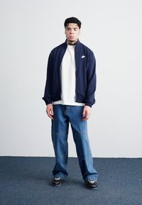 Casaco azul-marinho com fecho éclair, T-shirt branca, jeans azuis soltos e ténis pretos. Apresenta um logo da Nike no casaco, destacando o design desportivo.