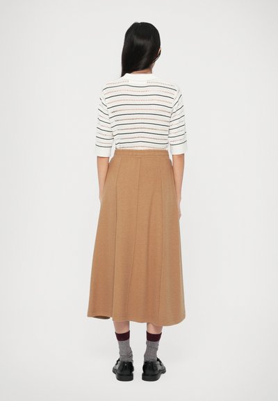 Donna con lunghi capelli scuri in piedi di spalle, che indossa una maglietta bianca a righe, una gonna midi color cammello, calzini grigi e scarpe nere.