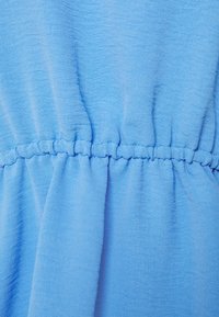 Světle modrá látka s mačkanou texturou má řasený pas, což vytváří jemný záhyb a hladké linie v designu oděvu.