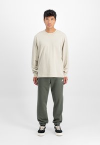 Béžové tričko s dlhým rukávom, vo voľnom strihu, v súlade s tmavo zelenými teplákmi s elastickými manžetami a malým logom. Čierne tenisky sú súčasťou.