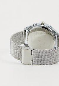 Silver stainless steel watch with a mesh band, featuring a polished closure and a smooth, reflective case back. Clear glass face visible.