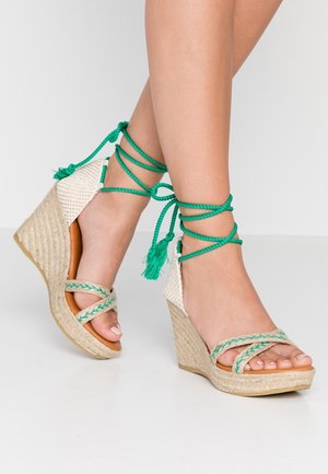 Kileformede espadriller sandal med beige flettede stropper og grønne ankelbind ud over fødder med lys hudtone og malede tånegle.