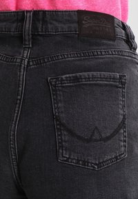 Jeans en denim noir avec une taille haute, dotés d'un design de poche arrière cousue et d'une étiquette en cuir brun avec logo au niveau de la taille.