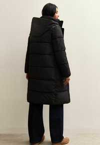 Cappotto lungo nero invernale con cappuccio, dotato di sezioni trapuntate, tasche laterali e una vestibilità rilassata, abbinato a jeans scuri e scarpe marroni.