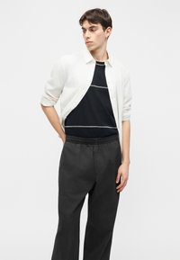 Fiatal férfi fehér gombos ingben, fekete, fehér varrással díszített pólóval, és bő, sötét szürke nadrággal, egyszerű háttér előtt.