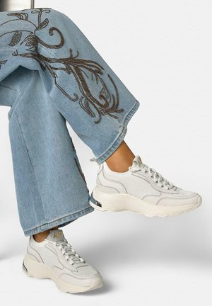 Jeans azzurri chiaro con ricami marroni abbinati a sneaker bianche chunky, con gambe incrociate su uno sfondo bianco.