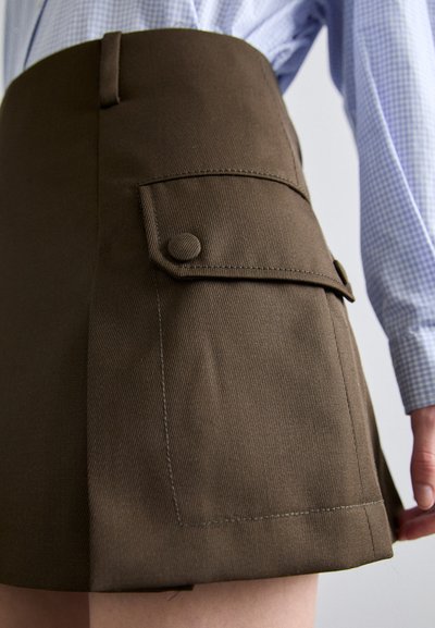 Καφέ ψηλόμεση φούστα με υφαντό ύφασμα, που διαθέτει πλευρική τσέπη με στρογγυλό κουμπί. Το τελείωμα είναι ίσιο με ορατές ραφές.