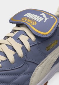 Κοντινό πλάνο ενός μπλε και λευκού αθλητικού παπουτσιού Puma King με μπεζ κορδόνια και καπάκι με την επιγραφή "Puma King" σε λευκά και χρυσά γράμματα.