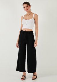 Biely rebrovaný crop top s čiernymi gombíkmi, v kombinácii s čiernymi širokými nohavicami a čiernymi sandálmi na opätku, s voľným strihom a sťahovacím pásom.