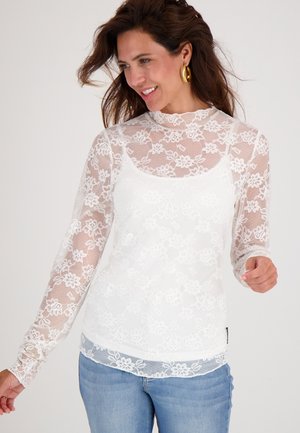 Witte kanten top met lange mouwen, versierd met bloemmotieven en een hoge kraag, gelaagd over een effen witte basis. Gecombineerd met blauwe jeans.