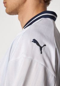 Weißes Sportshirt aus strukturiertem Stoff, mit vertikalen Streifen und einem kragen mit marineblauen Streifen. Verfügt über ein schwarzes Puma-Logo auf der linken Schulter.