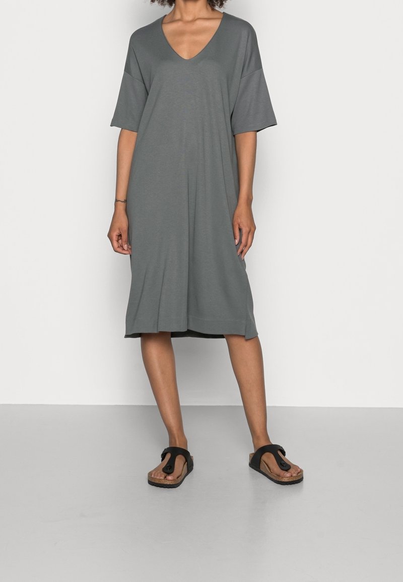 Robe grise à col en V avec des manches courtes, coupe décontractée, longueur genou, tissu doux et ourlet droit. Portée avec des tongs noires.