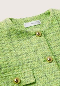 Gros plan sur une veste en tissu vert tissé avec des boutons dorés et un col, étiquetée MNG à l’intérieur de l’encolure.