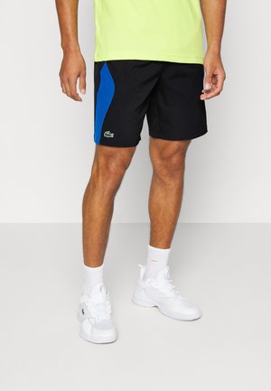 Pantaloni corti neri con pannello laterale blu, logo Lacoste in verde. Abbinati a una t-shirt giallo fluorescente e scarpe da ginnastica bianche.