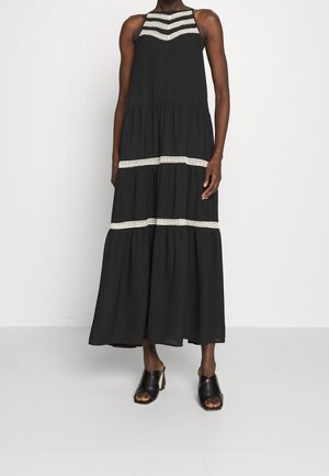 Frau trägt ein ärmelloses schwarzes Maxikleid mit weißen Spitzenstreifen über der Brust und an den Stufen, kombiniert mit offenen schwarzen Sandaletten mit Absatz.