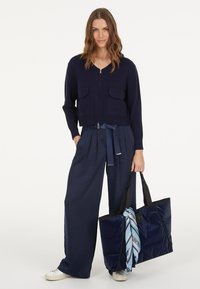 Marineblauer Strickcardigan mit Reißverschluss, zwei vorderen Taschen, kombiniert mit weit geschnittenen marineblauen Hosen, die mit einem Band zugeschnürt sind. Schwarze Tragetasche mit einem blauen Schal-Akzent.