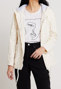 Person som bär en krämfärgad huva-jacka med ankermönster, en vit T-shirt med abstrakt ansiktsprint och högmidjade svarta byxor.