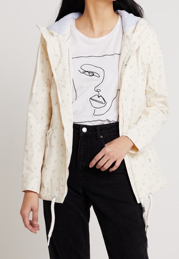 Person som bär en krämfärgad huva-jacka med ankermönster, en vit T-shirt med abstrakt ansiktsprint och högmidjade svarta byxor.