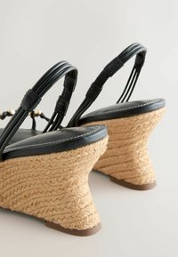 Sandales compensées noires à brides, avec talons en corde tressée et silhouette courbée, montrées de dos sur un fond clair.