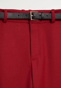Pantalon rouge avec couture avant et ceinture noire texturée avec une boucle rectangulaire dorée passée dans les passants de ceinture.