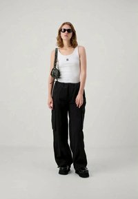 Bílý top bez rukávů s logem, černé cargo kalhoty, černé sluneční brýle a malá černá taška. Masivní černé boty doplňují vzhled. Minimalistický design.