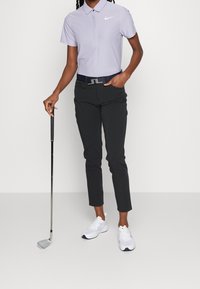 Světle fialová polo košile s límečkem a bílým logem, černé kalhoty a bílé tenisky. Drží golfovou hůl. Opasek má stříbrnou sponu.