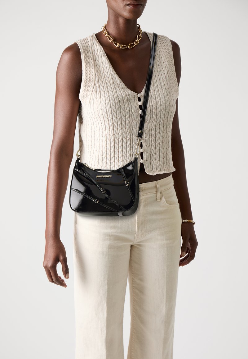 Black patent leather crossbody bag with a curved shape, featuring a gold logo, diagonal stitching, and a detachable strap.