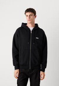 Svart zip-up hettegenser laget av bomullsblanding, med kengurulomme, justerbar snorhette og en liten logo på brystet.