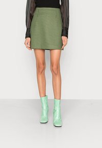 Modèle portant un pull noir à manches transparentes, une mini-jupe écossaise verte et des bottes cheville vert menthe, se tenant devant un fond blanc.