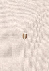 Tissu rose texturé avec un petit emblème de blason brodé au centre, présentant des bandes verticales beiges, blanches et brunes.