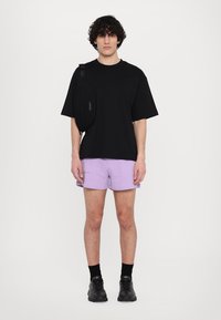 Črn oversize T-shirt, sivkast kratke hlače in črne superge. Torbica je čez eno ramo, stoji ob beli podlagi.