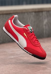 Raudona sportinė avalynė, pagaminta iš zomšos ir tinklelio medžiagų, su balta juosta, juodu padu ir ryškiu Puma logotipu ant liežuvėlio ir šono.