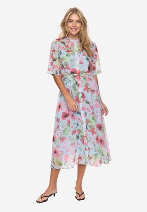 Kvinde smilende iført en midi-lang, knappet blomsterkjole med korte ærmer og sorte hælsandaler, stående mod en hvid baggrund.