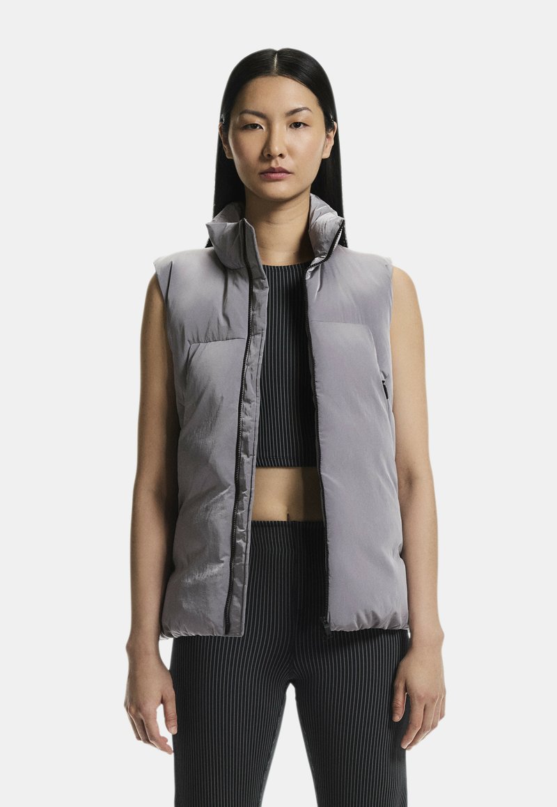 Grå polstret vest med højt krave, front lynlåslukning og to brystlommer. Bæres over en ribbet sort top og bukser med lodrette striber.