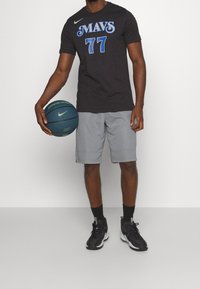 Czarny t-shirt z krótkim rękawem z niebieską grafiką "MAVS 77", połączony z szarymi szortami i czarnymi butami sportowymi, trzymający niebieską piłkę do koszykówki.