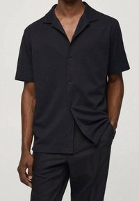 Uomo che indossa una camicia nera a maniche corte con colletto e pantaloni neri abbinati, in piedi con una mano in tasca.