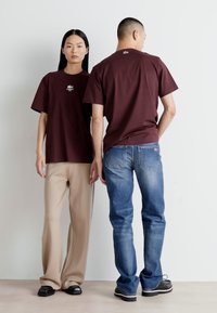 Vínové oversized trička s logem na hrudi, spojená s béžovými kalhotami a modrými džíny. Hladká bavlněná tkanina, volný střih, krátk�é rukávy.