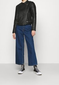 Giubbotto da biker in pelle nera con dettagli trapuntati sulle spalle, abbinato a jeans blu a gamba larga e sneakers nere alte.