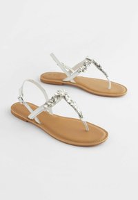 Witte platte sandalen met een metalen zilveren sieraadaccent over de banden, met een tan kleurige zool en een verstelbare enkelband.